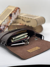 Load image into Gallery viewer, Brown leather wallet with money and cards on a white surface with a blurred newspaper in the background
